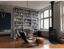 Wohnzimmer mit Holzboden, großem Bücherregal an der Wand, einer schwarzen modernen Kaminofenheizung, Sesseln und Ottoman. Große Fenster auf der rechten Seite lassen natürliches Licht herein, und ein Teppich liegt vor dem Kamin auf dem Boden.