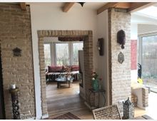 Offener Wohnraum mit teilweise freiliegenden Ziegelwänden, Holzbalkendecke und großen Fenstern, die viel Tageslicht einlassen. Im Vordergrund ein Tisch mit Stuhl, im Hintergrund ein kleiner Wohnbereich mit gemütlichen Sitzkissen und dekorativem Holztisch.