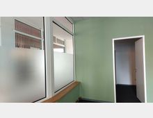 Innenraum mit einer grünen Wand, einer Glastrennwand mit Milchglasstreifen und einem offenen weißen Türrahmen, der in einen angrenzenden Raum führt. Der Boden ist mit dunklem Teppichboden bedeckt.