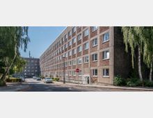 Mehrstöckiges Wohngebäude in Plattenbauweise mit rechteckigen Fenstern, entlang einer ruhigen Straße. Im Vordergrund stehen vereinzelt geparkte Autos, flankiert von Bäumen und Gehwegen.