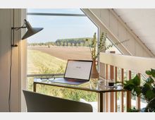 Schreibtisch in einem hellen Dachzimmer mit schrägem Fenster, Blick auf eine ländliche Landschaft mit Feldern und Bäumen. Auf dem Tisch stehen ein Laptop, eine Brille, eine Vase mit Zweigen und Blumen sowie eine Tasse Espresso.