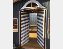 Eine geöffnete zweiflügelige Holztür mit diagonalen Streifen in Weiß und Blau führt in ein Gebäude mit Holzfußboden; im Inneren sind ein Kronleuchter und eine Glasbausteinwand sichtbar. Die Fassade des Gebäudes ist blau gestrichen, mit Fenstern und einem bogenförmigen Oberlicht über der Tür.