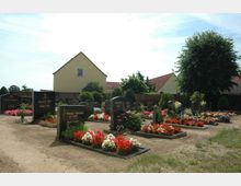 Ein kleiner Friedhof mit mehreren Grabsteinen, gepflegten Blumenbeeten und Kieswegen, umgeben von Bäumen und niedrigen Gebäuden mit roten Ziegeldächern. Im Hintergrund steht ein gelbes Wohnhaus.