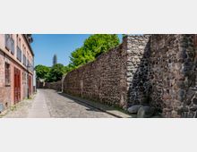 Gepflasterte Gasse mit einer historischen Steinmauer auf der rechten Seite und Gebäuden aus Backstein auf der linken Seite, umgeben von Bäumen und einem klaren blauen Himmel. Im Hintergrund ist ein Turm zu sehen, der teilweise von einem Baugerüst umhüllt ist.
