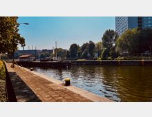Stadtkanal mit befestigtem Ufer, Bäumen im Hintergrund und einem Schleusenbereich; ein modernes Hochhaus ragt rechts aus der Begrünung empor.