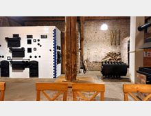 Innenraum einer rustikalen Backstube oder Werkstatt mit einer Wand aus weißen Fliesen mit schwarzen gusseisernen Ofentüren, einem zentralen Holzbalken, gestapeltem Brennholz im Hintergrund und einem schwarzen Ofen links. Die Umgebung ist von Backsteinwänden und Holzelementen geprägt.