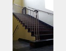 Eine Innenaufnahme einer Treppe mit dunklen Holzstufen und einem Metallgeländer mit gedrehten Stäben. Die Wand im Hintergrund ist hellgelb, und ein Handlauf aus Metall verläuft parallel zur Treppe.