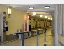 Eingangsbereich einer U-Bahn-Station mit hellem, gelbem Anstrich und Wandlampen. Im Hintergrund eine lange, beleuchtete Halle mit dekorativen Wandverkleidungen und Sitzbänken, vorne ein Geländer und ein öffentlicher Telefonautomat.