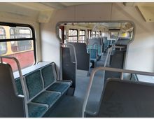 Innenraum eines regionalen Zuges mit einfachen, türkisfarben gepolsterten Sitzen auf beiden Seiten des Gangs. Große Fenster mit Blick auf den angrenzenden Zug und helle Wandverkleidungen definieren den Raum.