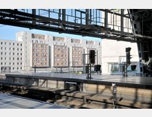 Bahnhof mit leeren Gleisen und Bahnsteigen im Vordergrund, einer Überdachung aus Metallstreben und Glaselementen sowie mehrstöckigen Wohngebäuden mit roten Backstein- und Betonfassaden im Hintergrund.