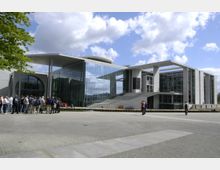 Modernes Regierungsgebäude mit großer Glasfassade und geometrischen Betonstrukturen, umgeben von einer gepflasterten Fläche. Im Vordergrund eine Gruppe von Menschen und vereinzelte Spaziergänger.