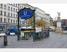Eingangsbereich des U-Bahnhofs Hermannplatz in Berlin mit einem blauen Schild mit der Aufschrift „Bhf. Hermannplatz“, umgeben von grünen Geländern. Im Hintergrund sind mehrstöckige Gebäude, Geschäfte wie McDonald's, eine Statue auf einem Sockel und eine gepflasterte Platzfläche mit Bäumen und parkenden Fahrrädern zu sehen.
