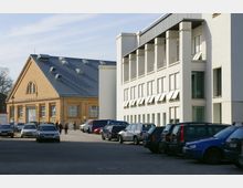 Mehrzweckgebäude mit einem gelben Backsteingebäude im traditionellen Stil links und einem weißen, modernen Bürogebäude mit rechteckigen Fenstern und Rollläden rechts; davor eine gepflasterte Fläche, die teilweise als Parkplatz genutzt wird.