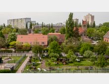 Ein ländlich gestalteter Bauernhof mit offenen Grünflächen, Traktoren und alten landwirtschaftlichen Geräten im Vordergrund, umgeben von Bäumen. Im Hintergrund sind Backsteinhäuser mit roten Dächern, eine Kirche mit Turm sowie moderne Wohnhochhäuser zu sehen.