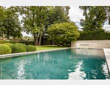 Privater Garten mit rechteckigem Pool, umgeben von gepflegten Hecken, Rasenflächen und großen Bäumen. Im Hintergrund eine beige Steinmauer mit integriertem Wasserspiel.