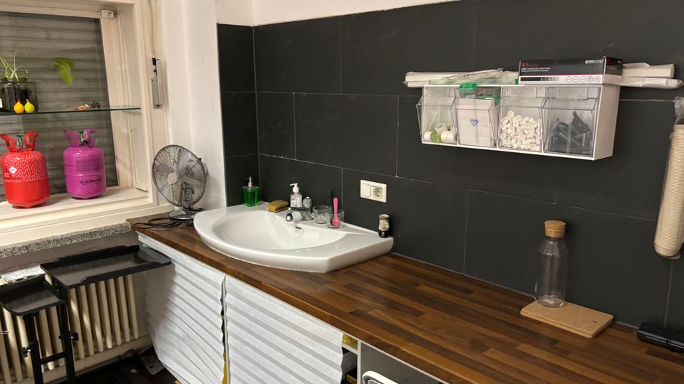 Kleiner Küchen- oder Arbeitsbereich mit langem Holzarbeitsplattenblock und eingelassenem Waschbecken vor einer dunklen Fliesenrückwand. Links befindet sich ein Fenster mit Ablage und Hockern, rechts hängen transparente Aufbewahrungsboxen und es stehen diverse Utensilien auf der Arbeitsfläche.