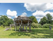 Holzspielanlage in Form eines Baumhauses auf Stelzen mit einer kleinen Rutsche, umgeben von einer grünen Wiese und großen Bäumen im Hintergrund unter einem bewölkten Himmel.