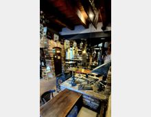 Rustikales Restaurantinterieur mit Backsteinwänden, Gewölbebögen und sichtbaren Holzbalken an der Decke. Mehrere Holztische und Stühle stehen entlang eines langen Raums, ergänzt durch hängende Lampen und dekorative Metallobjekte auf einem gemauerten Tresen.