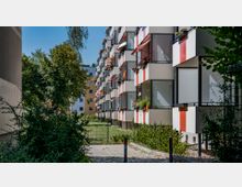 Moderne Wohngebäude mit Balkonen, die abwechselnd rot-weiße Fassadenelemente haben, dekoriert mit Pflanzen und Markisen. Im Vordergrund ist eine kleine Grünfläche mit Bäumen und Sträuchern zu sehen.