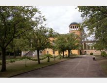 Historisches Gebäude mit gelber Fassade, einem Rundturm mit offener Säulenkonstruktion und Kuppel, umgeben von Bäumen und einem gepflegten Park. Ein gepflasterter Weg und eine Holzbank führen durch die Anlage.