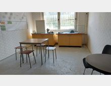 Ein einfacher Raum mit hellem Tapetenmuster, einem Tisch mit vier Holzstühlen und einer runden Sitzgelegenheit rechts. Vor einem Fenster mit Gitter befindet sich eine Holzkommode, darauf eine Glasvitrine mit ausgestellten Gegenständen.