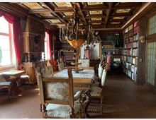 Bibliotheksraum mit historischem Interieur, darunter eine reich verzierte Holzdecke, hölzerne Bücherregale voller Bücher, ein großer Esstisch mit geschnitzten Stühlen und ein dekorativer Kronleuchter. An den Wänden befinden sich rote Vorhänge und diverse antike Verzierungen.