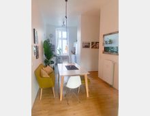 Essbereich in einer hellen Wohnung mit Holzfußboden, einem weißen Esstisch mit grüner Bank und weißen Stühlen. Im Hintergrund ist ein Fenster mit Vorhängen, eine Küchenecke und mehrere Zimmerpflanzen sichtbar.