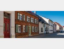 Straßenszene mit Reihenhäusern in einem Wohnviertel, darunter ein Backsteinbau mit grünen Fensterläden und daneben weiß verputzte Häuser mit dekorativen Blumenkästen; gepflasterte Straße und blauer Himmel im Hintergrund.