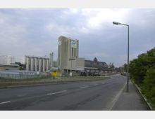 Industriegebiet mit mehrstöckigen Betongebäuden, darunter ein hohes Gebäude mit dem Logo „AMW“, und mehreren silbernen Silos. Im Vordergrund verläuft eine leere, asphaltierte Straße mit Laternen, rechts davon befindet sich Grünfläche.
