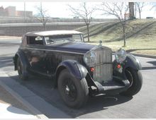Ein schwarzes Oldtimer-Cabriolet mit geschlossenem Verdeck steht auf einem Parkplatz am Straßenrand. Im Hintergrund sind kahle Bäume, ein leicht ansteigender Hügel und eine Straßenmauer zu sehen.