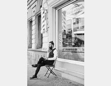 Ein schwarz-weiß-Bild zeigt einen Mann in formeller Kleidung, der auf einem Stuhl vor einem Friseursalon mit der Aufschrift „Küçük Istanbul Barber Shop“ sitzt. Neben der Eingangstür hängt eine klassische rot-weiß gestreifte Friseur-Stange, und auf dem Fenster sind weitere Friseur-Elemente und Schriftzüge sichtbar.