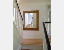 Eine lichte, helle Treppenhaus-Szene mit weißen Geländern und einem Holzfußboden. An einer Wand hängt ein großer, vergoldeter Spiegel in einem reich verzierten Rahmen, der den Treppenverlauf reflektiert.