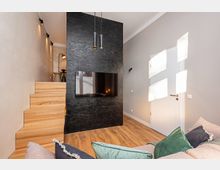 Modernes Wohnzimmer mit Holzfußboden und minimalistischer Einrichtung, dominiert von einer schwarzen Akzentwand mit integriertem Flachbildfernseher und hängenden Pendelleuchten. Links führt eine helle Holztreppe zu einer erhöhten Nische, rechts befindet sich eine weiße Tür in einer grauen Wand mit einfallendem Sonnenlicht.