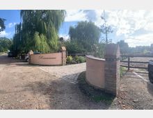 Eingangsbereich eines Anwesens mit einer geschwungenen Mauer aus Putz und Backstein, auf der der Schriftzug „Rosencarree“ steht. Im Hintergrund sind ein Kiesweg, ein Zaun, parkende Autos sowie Bäume und Sträucher zu sehen.