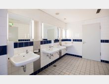 Öffentlicher Waschraum mit weißen Wandfliesen und blauen Fliesendetails. Drei weiße Waschbecken mit Spiegeln und Seifenspendern an der Wand, plus eine geschlossene Tür im Hintergrund.