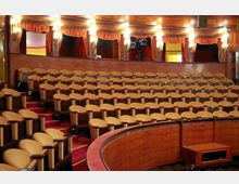 Ein klassischer Zuschauerraum in einem Theater oder Opernhaus mit gepolsterten, beigefarbenen Sitzreihen und großzügiger Holzvertäfelung. Der Raum ist mit roten Vorhängen und eleganten Wandleuchten dekoriert.