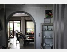 Wohnzimmeransicht mit einem durch einen Rundbogen sichtbaren offenen Bereich, der zu einem Raum mit einer großen Glas-Schiebetür führt. Der Vordergrund zeigt einen grauen Schrank mit Geschirr und dekorativen Elementen, im Hintergrund Ledermöbel, ein Couchtisch und ein Blick ins Freie.