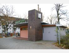 Backsteingebäude mit abgerundeten Elementen und einem Schriftzug "Imbissbüdchen" auf dem Dach, daneben ein kleiner Außenbereich mit Fahrradständer und Sitzgelegenheiten; im Hintergrund sind Bäume und Wohnhäuser zu sehen.