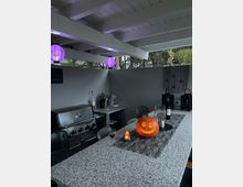 Überdachte Outdoor-Küche mit grauen Arbeitsflächen, einem Edelstahlgrill, lila Laternen an der Decke und dekorativen Elementen wie einem beleuchteten Kürbis und Kerzen auf dem Tisch. Im Hintergrund befinden sich graue Wände und Bäume außerhalb der offenen Seiten.