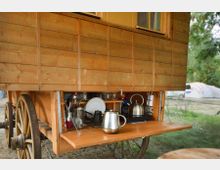 Holzwagen mit aufklappbarer Seitenwand, die eine kleine Außenküche zeigt; enthalten sind Küchenutensilien, Geschirr und ein Wasserkocher. Im Hintergrund sind Zelte und Bäume auf einer Wiese zu sehen.