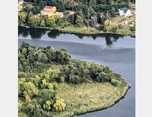 Luftaufnahme einer Flusslandschaft mit einer bewaldeten Insel im Vordergrund und mehreren Wohnhäusern am gegenüberliegenden Ufer, umgeben von vielen Bäumen. Der Fluss spiegelt den Himmel und gibt der Szene eine ruhige Atmosphäre.