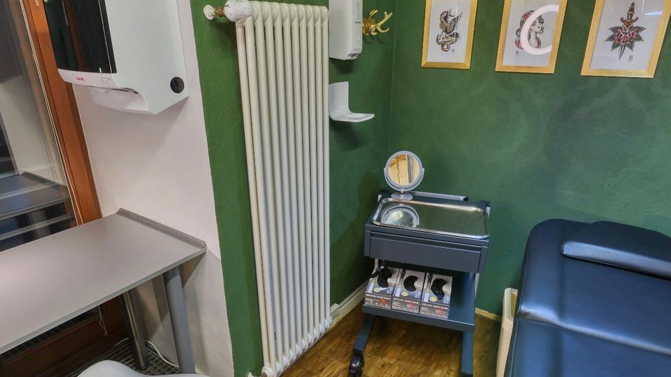 Kleiner Behandlungsraum mit dunkelgrünen Wänden, Holzfußboden und einer schwarzen Untersuchungsliege neben einem rollbaren Metalltisch mit Ablage. An den Wänden hängen gerahmte Motive und Regale mit Deko, zudem sind ein Heizkörper, ein Waschmittelspender und ein Hocker am Arbeitstisch zu sehen.
