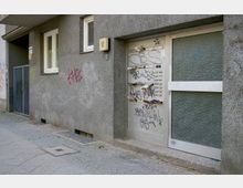 Fassade eines Wohngebäudes mit grauem Putz, einer Haustür aus Metall und Glas sowie einem Briefkastensystem, das mit Graffiti bemalt ist. Der Gehweg aus Pflastersteinen ist stellenweise mit Laub bedeckt.