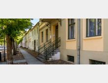 Straßenzug mit alten, hell gestrichenen Wohnhäusern und hohen Fenstern, davor ein Gehweg entlang einer Baumallee. Im Vordergrund führt eine Treppe mit schmiedeeisernem Geländer zu einem Hauseingang; Kopfsteinpflaster und fallendes Laub säumen den Rand.
