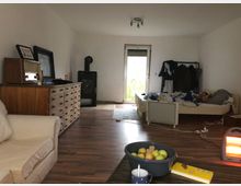 Wohnzimmer mit einfacher Einrichtung, darunter ein Sofa, ein Holzschrank mit Schubladen, ein Kaminofen und ein Bett. Im Vordergrund sind ein elektrischer Heizlüfter, eine Schüssel mit Obst und eine Tasse auf einem niedrigen Tisch zu sehen; im Hintergrund ist eine Tür zum Außenbereich.