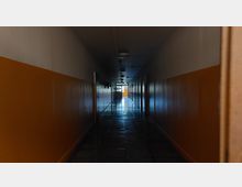 Langer, schmaler Flur in einem Gebäude mit orangefarbenen und weißen Wänden, gefliesten Böden und mehreren geschlossenen Türen auf beiden Seiten. Am Ende des Flurs ist ein Fenster mit einfallendem Tageslicht sichtbar.