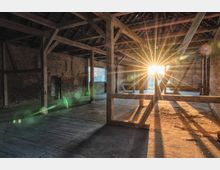 Eine leere Halle mit Holzstützbalken und sichtbarem Mauerwerk, durch deren geöffnete Tür die tief stehende Sonne hereinscheint und Lichtstrahlen in den Raum wirft. Der Boden ist aus Beton, und die Decke besteht aus einem sichtbaren Dachstuhl.