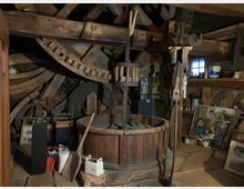 Innenraum einer historischen Schmiede oder Werkstatt mit Holzkonstruktionen, großen Zahnrädern und einem zentralen hölzernen Bottich. Im Hintergrund sind weitere Werkzeuge, Geräte und dekorative Objekte wie Gemälde und Keramik sichtbar.
