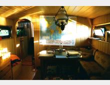 Innenraum eines kleinen, holzvertäfelten Wohnbereichs mit einer Sitzbank und einem Tisch, auf dem Bücher und Gegenstände liegen. Eine Weltkarte hängt an der Wand, während Licht durch seitliche Fenster hereinfällt und einen Kronleuchter beleuchtet.