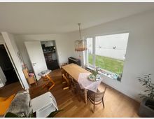 Esszimmer in einer modern eingerichteten Wohnung mit Holzboden, einem großen Holztisch samt Stühlen und einer dekorierten Tischdecke. Das Zimmer hat eine große Fensterfront mit Blick auf eine Wiese und weiße Wände, an der Wand sind Vogelsilhouetten angebracht.